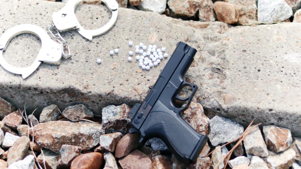 A BB gun is on concrete next to a small pile of white BBs, and a pair of handcuffs, indicating the potential for weapon charges in New York for these and similar weapons.