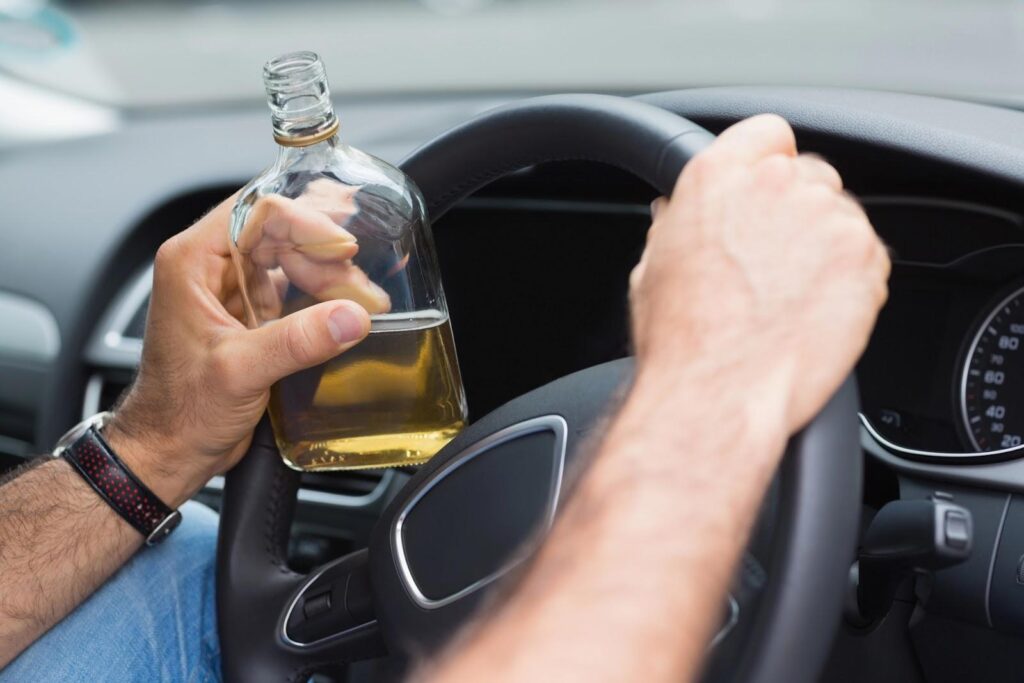 A man’s hands grip a steering wheel, with a bottle of alcohol in his left hand just before a DUI arrest that may affect his immigration application.