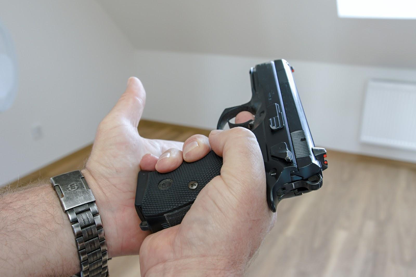 White male hands with a metal watch on the left wrist hold a pistol inside their home, and the individual could face weapon charges if they take the pistol outside.